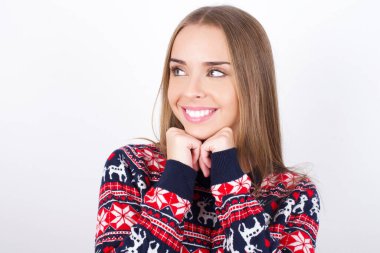 Tatmin olmuş, beyaz arka planda Noel süveteri giyen beyaz tenli genç bir kız çenesine dokunuyor, iki eliyle gülümsüyor, sevgilisiyle mutlu bir gün geçiriyor.