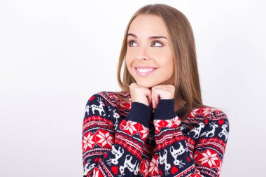 Beyaz arka planda Noel süveteri giyen mutlu beyaz kız harika bir şey olacağını umuyor, mutlu bir şekilde yan yana bakıyor, el ele tutuşuyor, mutlu bir yüz ifadesi var..