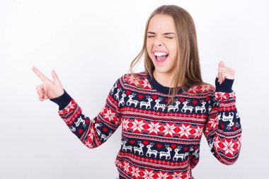 Beyaz arka planda Noel süveteri giyen neşeli beyaz kız fotokopi alanı reklamını göstererek şansı kutluyor.