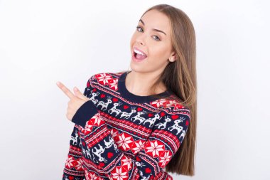 Beyaz arka planda Noel süveteri giyen beyaz tenli genç beyaz kız fotokopi alanını gösteren neşeli bir gösteri sunuyor.