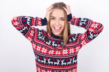 Neşeli, neşeli, beyaz arka planda Noel süveteri giyen Kafkasyalı genç kız harika haberler aldıktan sonra ellerini havaya kaldırarak tepki veriyor..
