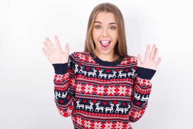 Beyaz arka planda Noel süveteri giyen Kafkasyalı genç kız harika bir sonuç elde ettikten sonra ağzını ve kollarını açar.