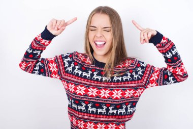 Beyaz arka planda Noel süveteri giyen çılgın beyaz bir kızın fotoğrafı. Çığlık atıyor ve parmakları saçları kapalı gözleri işaret ediyor.