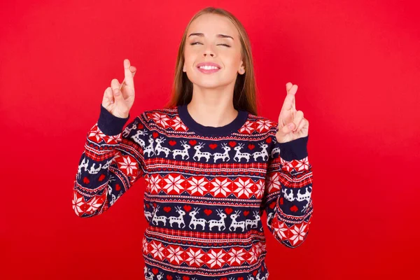 身穿圣诞毛衣的年轻高加索女孩满怀希望 叉着手指 相信好运 面带微笑 人与愿望概念 — 图库照片