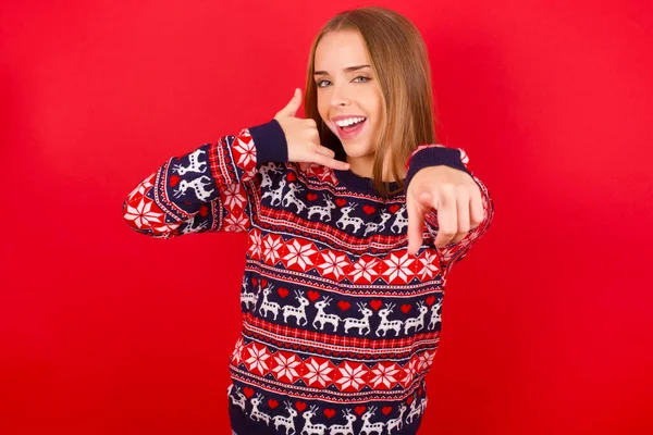 Kafkasyalı genç bir kız Noel süveteri giyiyor, neşeli bir şekilde gülümsüyor ve kamerayı işaret ediyor, daha sonra seni arıyor, telefonla konuşuyor.