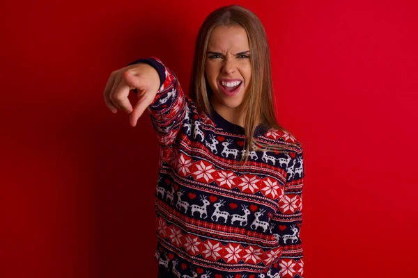 Kırmızı arka planda Noel süveteri giyen genç beyaz kız kameraya kızgın ve sinirli bir şekilde sizinle kavga etmeye hazır olduğunu gösteriyor..