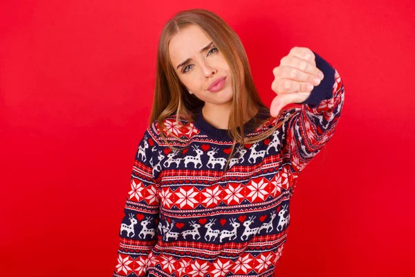Kırmızı arka planda Noel süveteri giyen genç beyaz bir kız kızgın, sinirli, hayal kırıklığına uğramış ya da hoşnutsuz, ciddi bir bakışla başparmaklarını göstermiyor.
