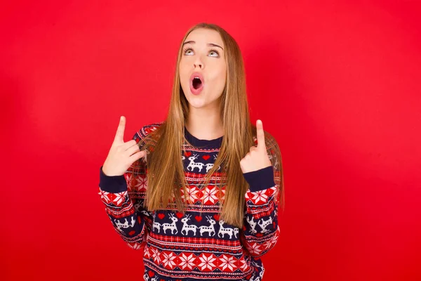 年轻的高加索女孩穿着红色背景的圣诞毛衣 惊讶地抬起头 用手指和胳膊指指着 — 图库照片