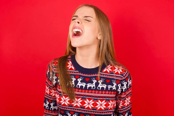 Kafkasyalı genç bir kız kırmızı arka planda Noel süveteri giyiyor kızgın ve öfkeli çığlıklar atıyor, öfkeyle bağırıyor. Öfke ve agresif kavram.