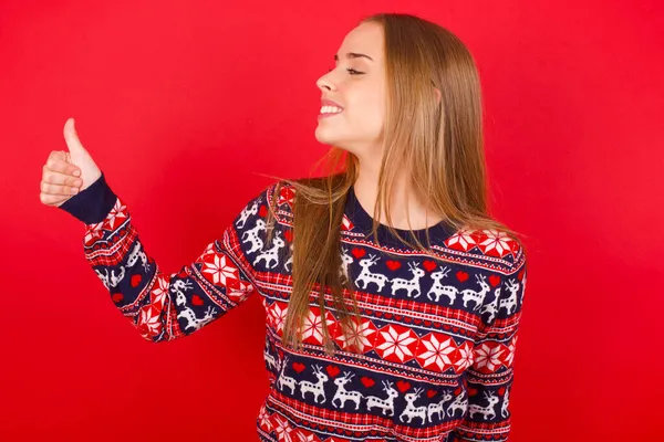 年轻的高加索女孩穿着红色背景的圣诞毛衣看起来很自豪 微笑着向旁边做着竖起大拇指的手势 干得好 — 图库照片