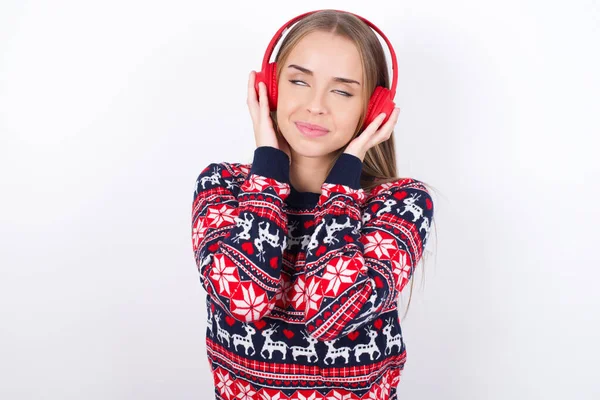 快乐的年轻高加索女孩穿着白色背景的圣诞毛衣 喜欢听悦耳的旋律 手握立体声耳机 闭上眼睛 与音乐共度业余时间 — 图库照片