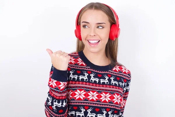 年轻的高加索女孩穿着白色背景的圣诞毛衣 通过无线耳机听音频 然后用大拇指将广告复制到其他地方 — 图库照片