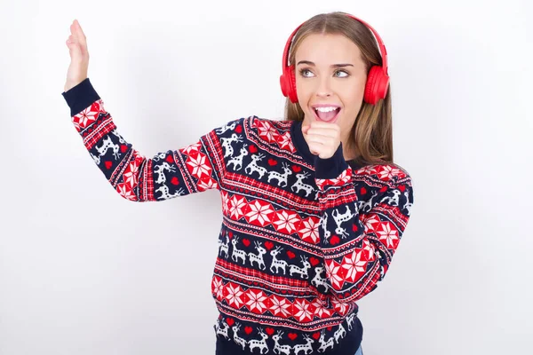 快乐的年轻高加索女孩穿着白色背景的圣诞毛衣唱着最喜欢的歌 手紧紧抓住嘴 好像话筒戴着无线耳机 听着音乐 — 图库照片