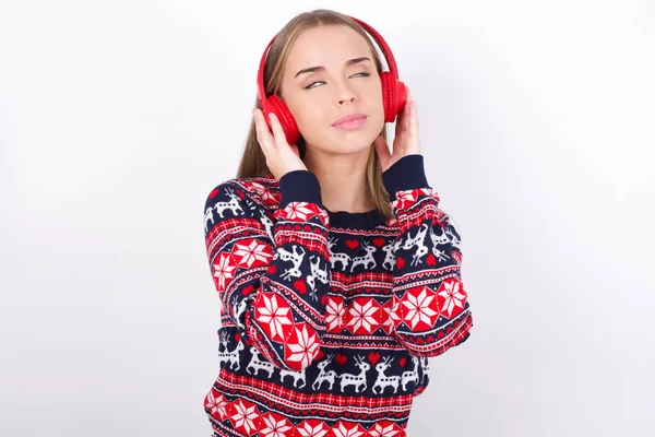 Mladá Běloška Nosí Vánoční Svetry Bílém Pozadí Sluchátky Hlavě Poslouchá — Stock fotografie