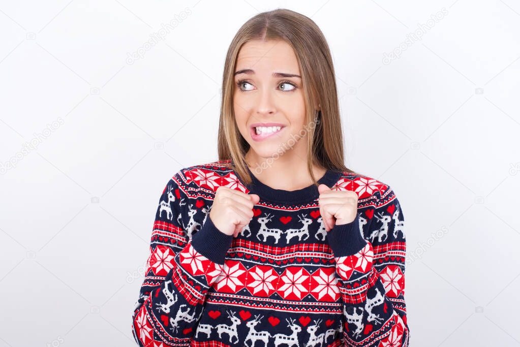 Young caucasian girl wearing christmas sweaters on white background clenches fists and awaits for something nice happened looks away bites lips and waits announcement of results
