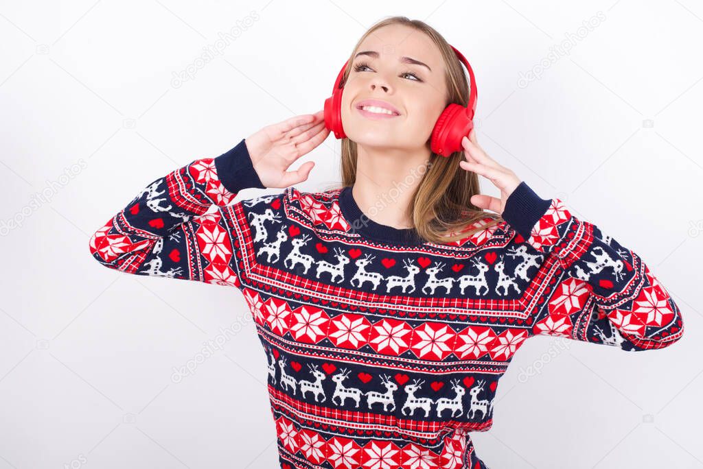 Joyful Young caucasian girl wearing christmas sweaters on white background sings song keeps hand near mouth as if microphone listens favorite playlist via headphones
