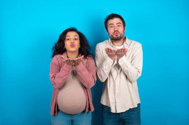 Genç bir çift mavi arka planda bir bebek bekliyor. Öpücük yolluyor, dudaklarını büzüyor ve hava öpücüğü vermek için avuç içi tutuyor..