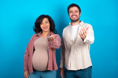 Mavi arka planda bebek bekleyen genç bir çift mutlu bir şekilde gülümseyerek yardım eli uzatıyor ve kabul ediyor..