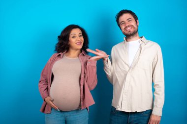 Genç bir çift mavi arka planda bir bebek bekliyor. Gülümsüyor, mutlu bir şekilde sunuyor ve avuç içi kameraya bakıyor..