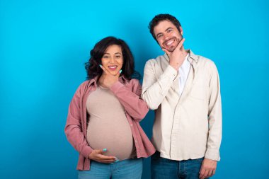 young couple expecting a baby standing against blue background looking confident at the camera smiling with crossed arms and hand raised on chin. Thinking positive.