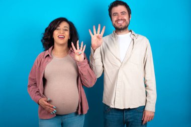 Genç çift mavi arka planda duran bir bebek bekliyor. Dört numaralı parmaklarıyla işaret ediyor ve gülümsüyor. Kendinden emin ve mutlu..