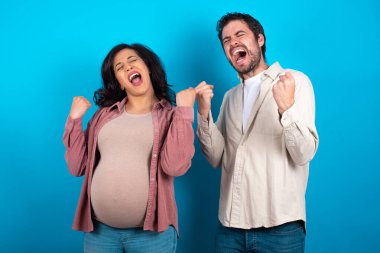 Mavi arka planda duran genç bir çift kolları havada ve gözleri kapalı bir şekilde başarıyı kutluyor ve şaşırıyor. Kazanan konsept.