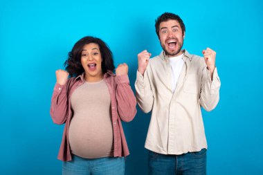 Mavi arka planda duran genç bir çift kollarını kaldırıp gözleri açık bir şekilde başarıyı kutluyor ve şaşırıyor. Kazanan konsept.