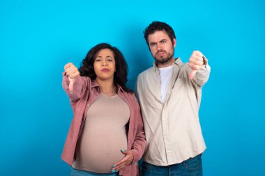Genç bir çift mavi arka planda bir bebek bekliyor kızgın, sinirli, hayal kırıklığına uğramış ya da hoşnutsuz hissediyorlar, ciddi bir bakışla başparmaklarını gösteriyorlar.