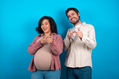 Genç bir çift mavi arka planda durup mutlu ve komik yüzlü parmakları kameraya gösteren bir bebek bekliyor. İyi enerji ve titreşimler.