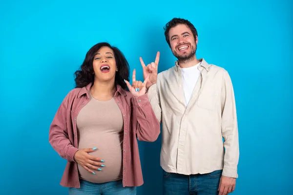 Young Couple Expecting Baby Standing Blue Background Doing Rock Gesture — Stock Photo, Image