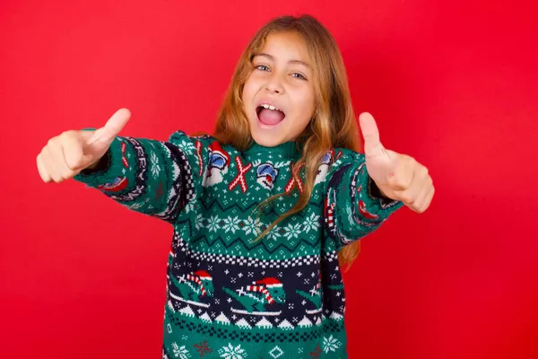 面白いブルネットの子供の女の子は赤い背景の上にニットセータークリスマスに親指で積極的なジェスチャーを作ります笑顔と成功のために幸せ カメラを見て勝者のジェスチャー — ストック写真