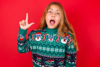 Kırmızı arka planda örgü süveter giyen hoş görünümlü esmer kız çocuğunun akıllı bir ifadesi var, bir parmağını kaldırıyor, önemli bir şeyi söylemeyi unutmaması gerektiğini hatırlıyor..