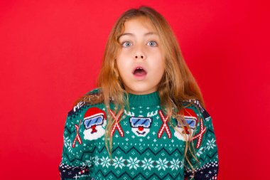 Kırmızı arka planda örgü süveter giyen, duygusal ve çekici esmer kız ağzı açık, büyük bir şaşkınlık ve korku içinde kameraya bakıyor. Beklenmedik haberler ve insani tepkiler.