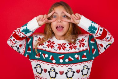 Kırmızı arka planda örülü süveter giyen neşeli, pozitif esmer kız, ağzının kenarında V işareti gösteriyor.