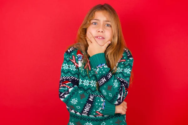 黑发小女孩 穿着针织毛衣 背着红色背景的圣诞节 双手捂着嘴 被什么东西或某人咬着钉子吓着了 — 图库照片