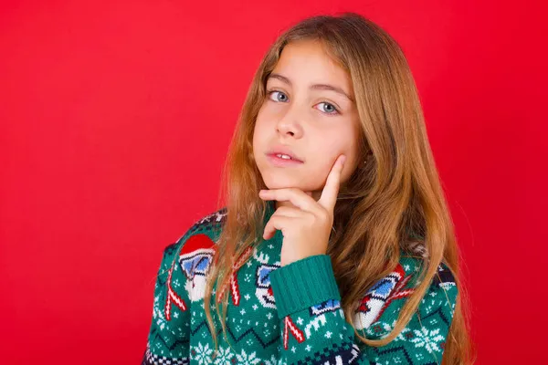 慎重に成功したブルネットの子供の女の子は 赤い背景の上にニットセーターのクリスマスに 白い歯を喜んで輝く頭を傾ける顎の視線カメラに触れています 歯科医療の概念 — ストック写真