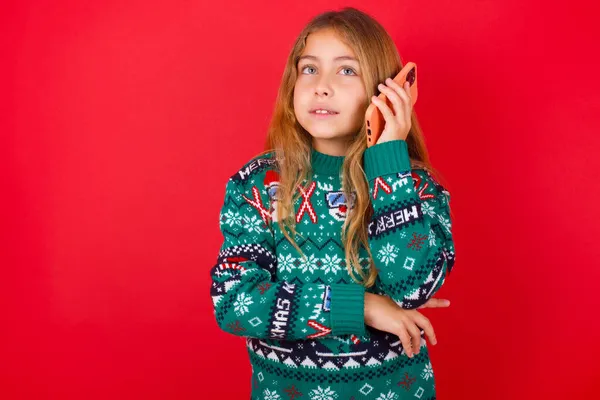 携帯電話で話して赤い背景の上にニットセーターのクリスマスで笑顔面白いブルネットの子供の女の子の肖像画 ビジネス コミュニケーションの概念 — ストック写真