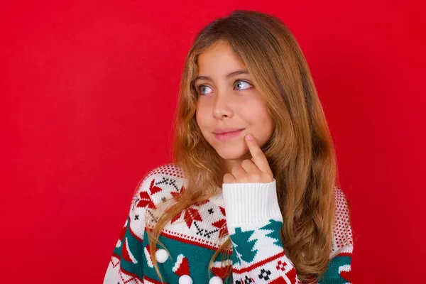 穿着针织毛衣 背景为红色的圣诞节的可爱的 梦幻般的黑发小女孩把手指放在嘴边看着旁边复制的空间 — 图库照片