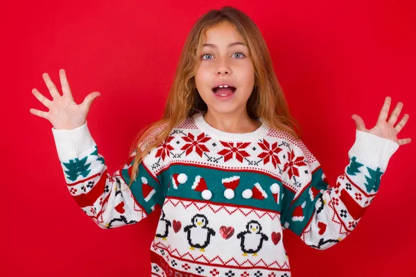 红色背景下 穿着针织毛衣圣诞节的快乐的 积极的黑发小女孩在取得重大成果后张开了嘴和胳膊 — 图库照片