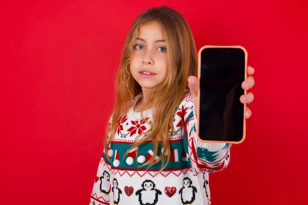 魅力的な可愛いですブルネット子供女の子でニットセータークリスマス以上赤背景保持現代的なデバイス ショー黒画面スマートフォン — ストック写真