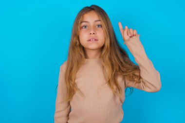 Mavi arka planda süveter giyen esmer kız bir numarayı gösterip işaret ederken kendinden emin ve mutlu bir şekilde gülümsüyor..