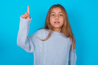 Mavi arka planda mavi örülü süveter giyen esmer kız parmağıyla işaret ediyor ve dahiyane düşüncelerden esinlenerek bakıyor, iyi bir fikir işareti gösteriyor, zekice bir çözümü var.