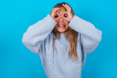 Mavi arka planda mavi örülü süveter giyen esmer kız dürbün gibi dil çıkartıyor, gözler parmaklarından bakıyor. Çılgın ifade.