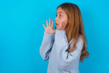 brunette kid girl wearing blue knitted sweater over blue background shouts loud, keeps eyes opened and hands tense. clipart