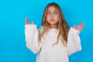 Öfkeli esmer kız beyaz örgü süveter giyiyor mavi arka plan jestleri şaşkınlık içinde, hoşnutsuzca kaşlarını çatıyor..