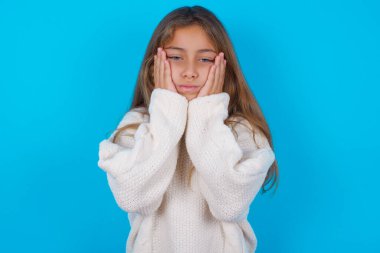 Esmer kız mavi arka planda beyaz örülü süveter giyiyor. Yüzü, depresyonu ve üzüntüyü örten eller, sorun yüzünden üzgün ve sinirli.