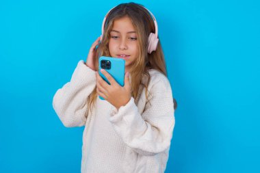 Mavi arka planda beyaz örgü süveter giyen mutlu esmer kız akıllı telefon ekranında odaklanırken iyi hissediyor. İnsanlar, teknoloji, yaşam tarzı