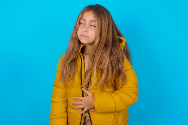 Brunette Kid Girl Wearing Yellow Jacket Blue Background Touches Tummy — Stock Photo, Image