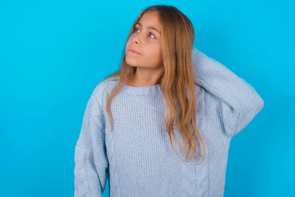 青い背景に青いニットセーターを着たブルネットの子供の女の子は混乱し 質問について疑問に思っています 疑いもなく 頭の上で手で考えて 包括的概念 — ストック写真