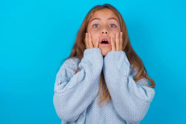 青い背景に青いニットセーターを身に着けている洗練されたブルネットの子供の女の子は興奮とスリルを表現し 顎を落とし続け 頬に手を出し 目が出てきました — ストック写真